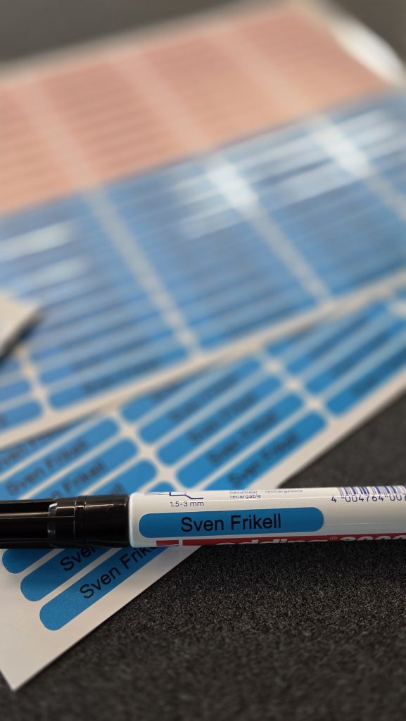 Namensetikett bzw. Namensaufkleber auf einem Stift. Im Hintergrund mehrere versandfertige Bögen im A4-Format aus der automatisierten Produktion kleinformatiger Namensetiketten.