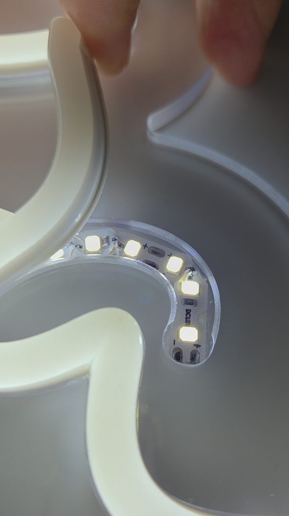 In einer Fräsnut liegt das LED-Band, welches im Bogen des Buchstabens gestaucht ist. Eine Hand hält das Silikonband nach oben. LED- und Silikonband gehören zum Produkt LED Flex Neon Generation 2 von Farben-Frikell | WTB.