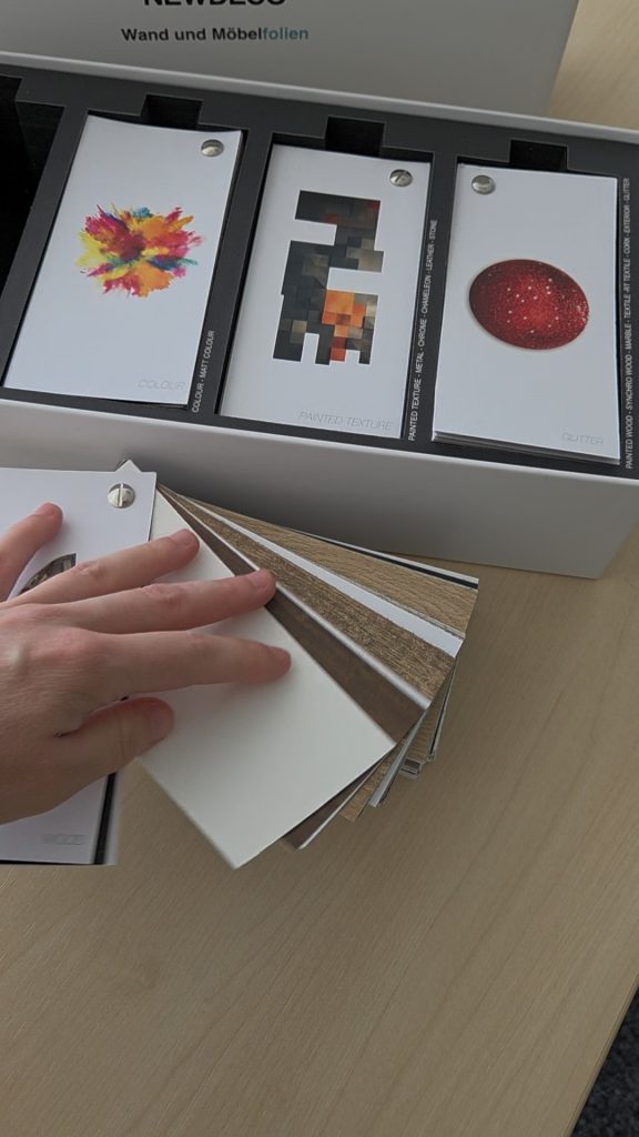 Musterfächer aus der Musterbox der Möbelfolien von New Deco. Eine Hand blättert durch den Fächer mit Holzdekor.